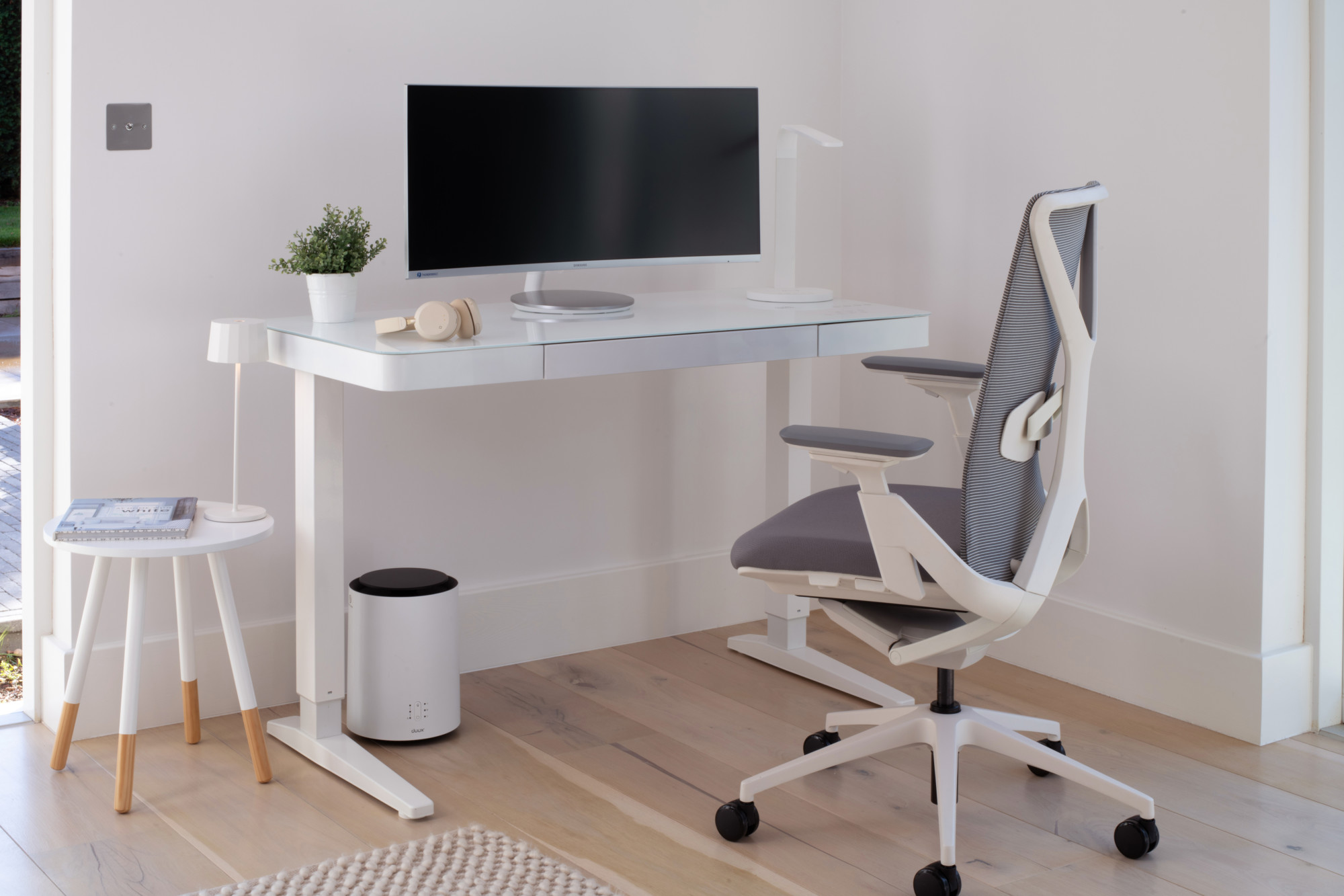 White frame and glass top standing desk in a home office environment paired with a white and grey office chair. 
