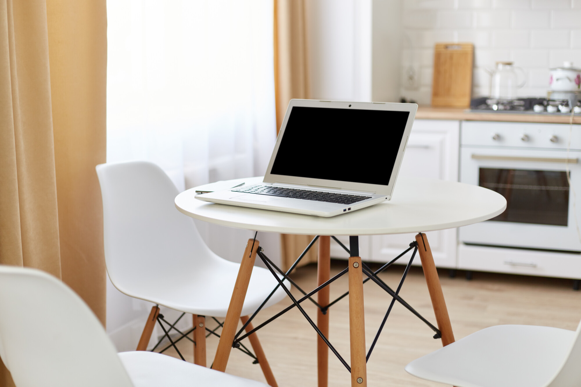 Round white table with laptop with blank screen for advertisement and smart phone on it, workspace for freelancer at home in light kitchen room near window.
